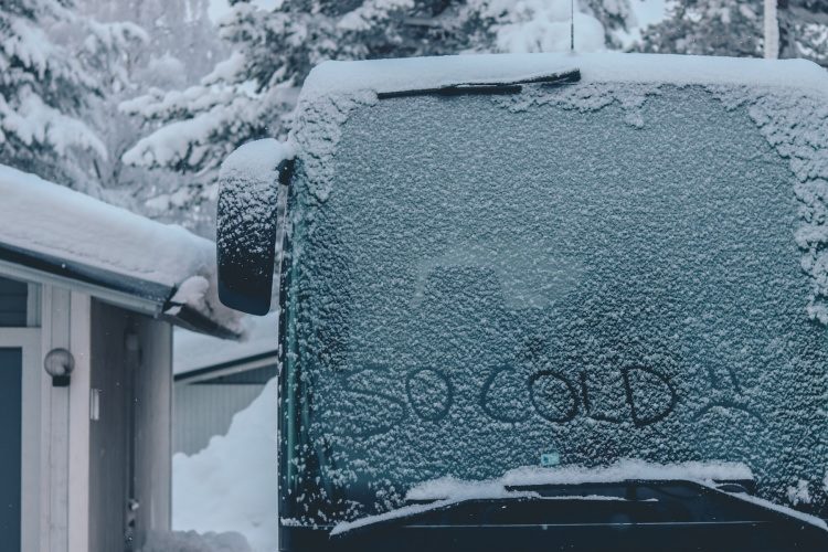Van with SO COLD written in frost on windshield beside white and blue residential garage