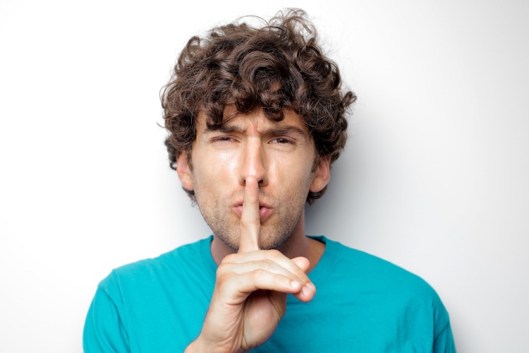 Man with brown hair holding finger to lips in shushing motion