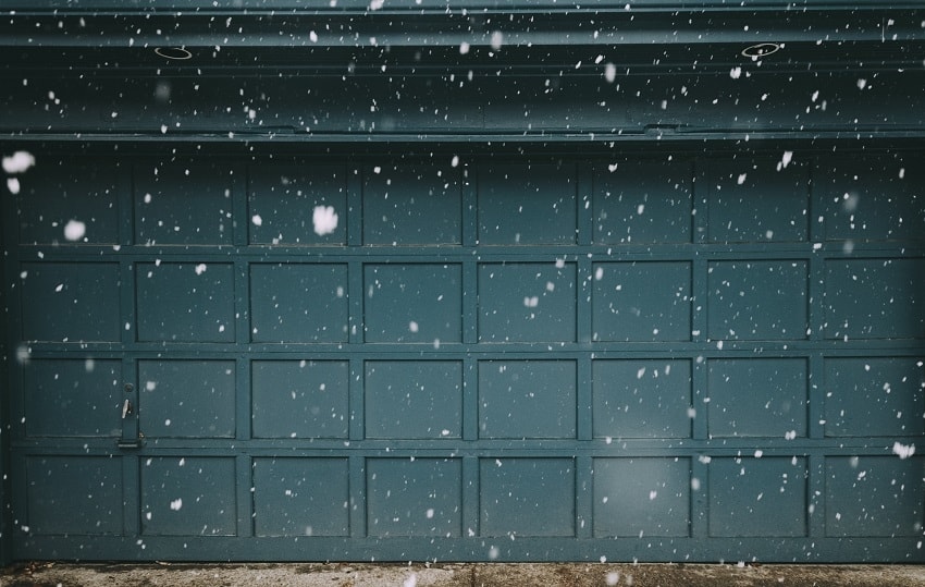 Dark teal paneled two-car garage door through flakes of falling snow in winter season