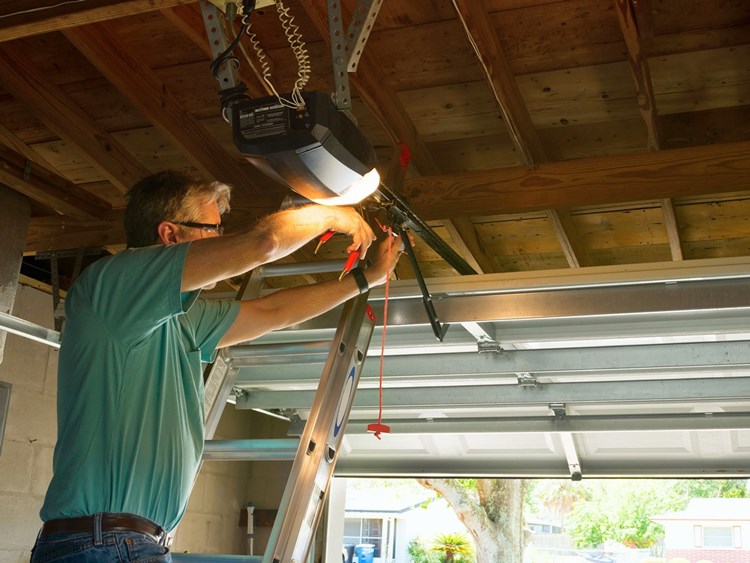 Homeowner on step ladder repairing garage door opener inside residential garage